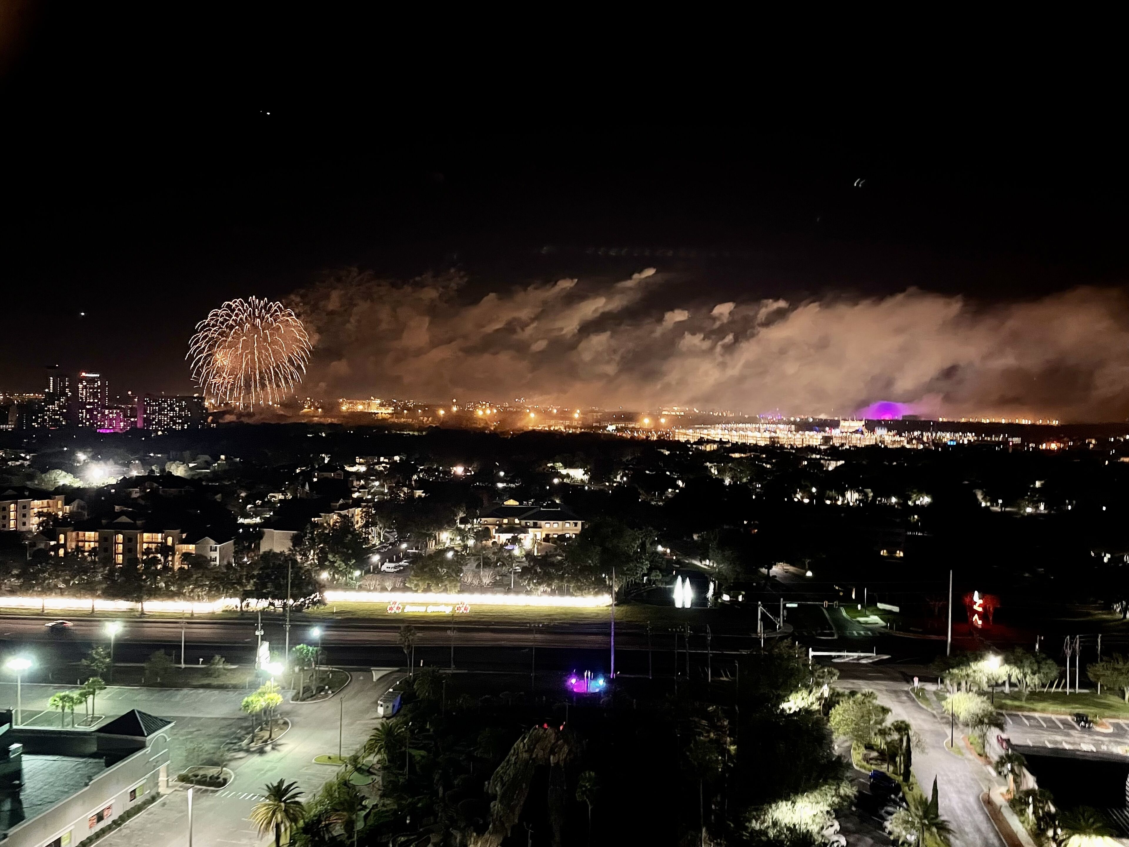 Une vue nocturne d’une ville, avec un feu d’artifice, des bâtiments illuminés et un parking.