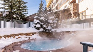 Un jacuzzi rodeado de árboles cubiertos de nieve y un muro de piedra, con un edificio al fondo.