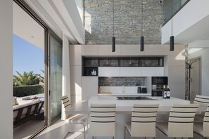 A modern kitchen with a stone accent wall, a dining area with striped chairs, and a view of palm trees outside. - Impeccable design, total privacy & heated pool with sea views (Rethymno)
