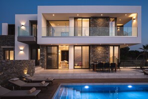 Une maison moderne à deux étages, dotée d’une piscine, de balustrades en verre et d’éléments décoratifs en pierre.