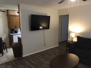 A living room with a flat-screen TV mounted on the wall, a dining area with wooden cabinets, and a kitchen area with a dishwasher.