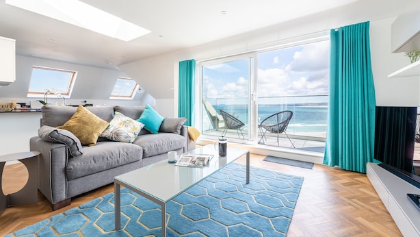 A modern living room with a grey sofa, a glass coffee table, and a view of the sea through large windows.