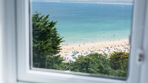 A beach view through a window, featuring a sandy beach, clear blue water, and green foliage.