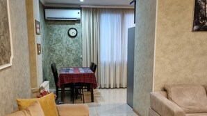 A living room with a beige sofa, yellow cushions, a dining area with a red and black checkered tablecloth, and a clock on the wall. - Niniko's Old Tbilisi Historical House. (Tbilisi)