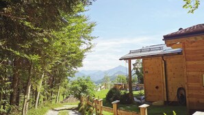 A wooden cabin with a gravel path leading to it, surrounded by lush greenery and mountains in the background.