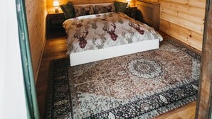 A cozy wooden cabin bedroom with a bed, two bedside lamps, a window with curtains, a framed picture on the wall, and a patterned rug.