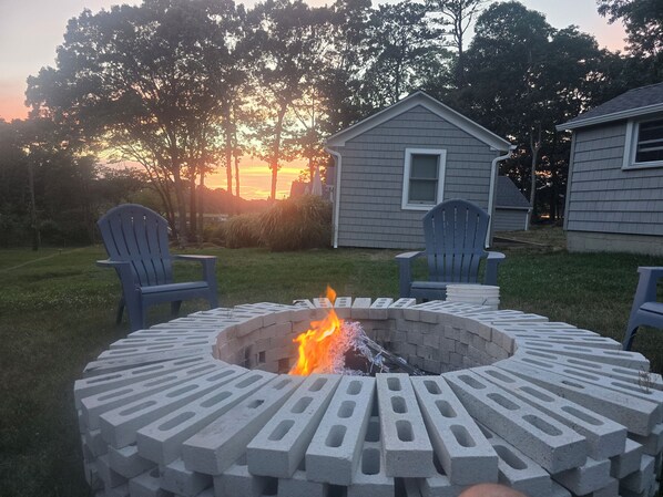 A circular fire pit with a fire burning, surrounded by chairs, set against a house and trees during sunset.