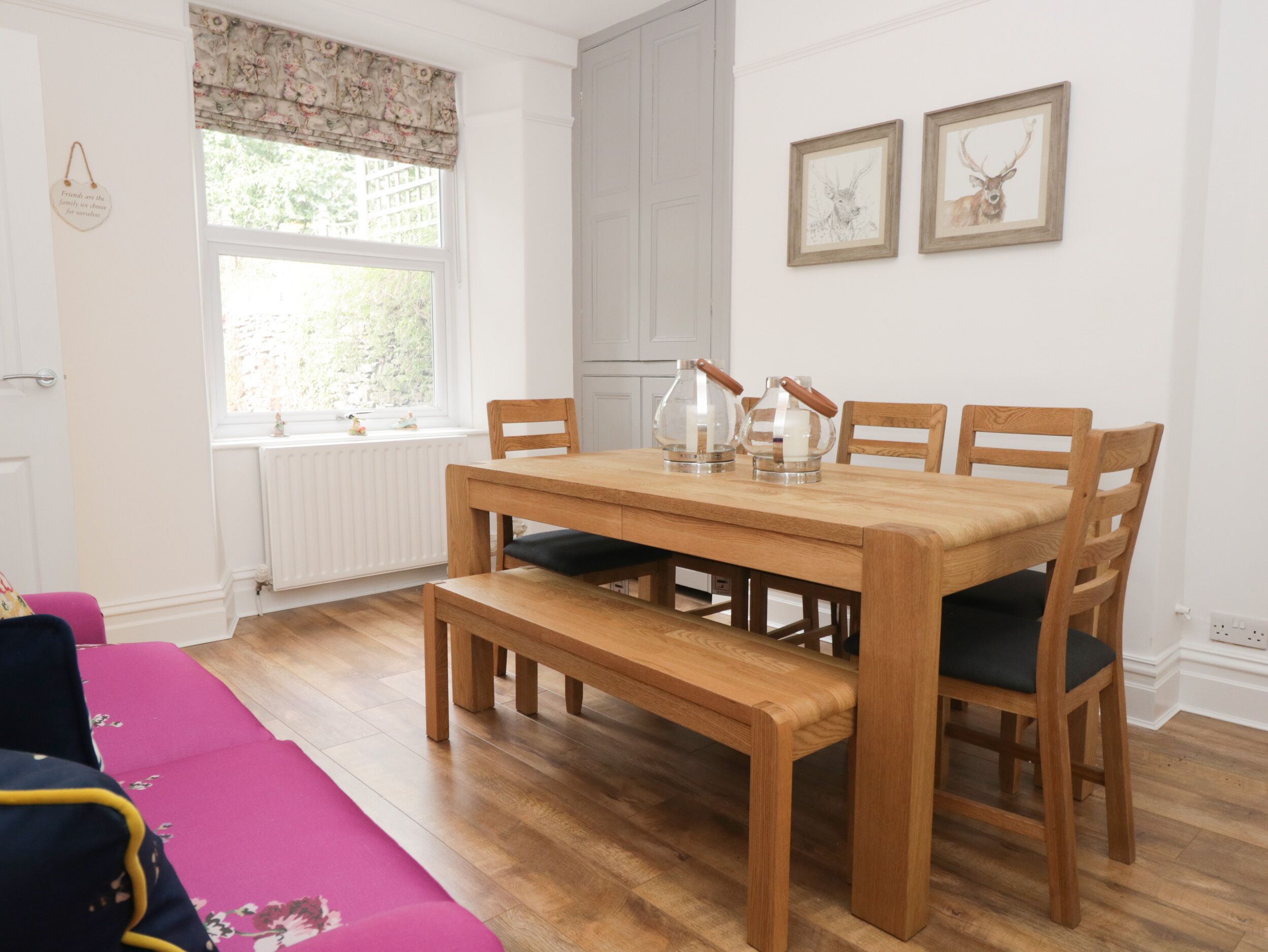 A dining area with a wooden table and chairs, a bench, and a window with a floral valance. There are two framed pictures on the wall.