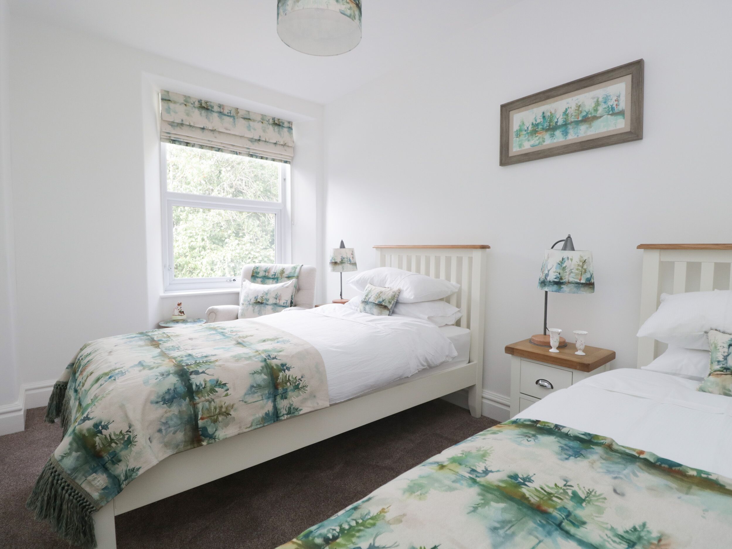 A bedroom with two beds, a window with a view of greenery, a bedside table with a lamp, and a framed picture on the wall.