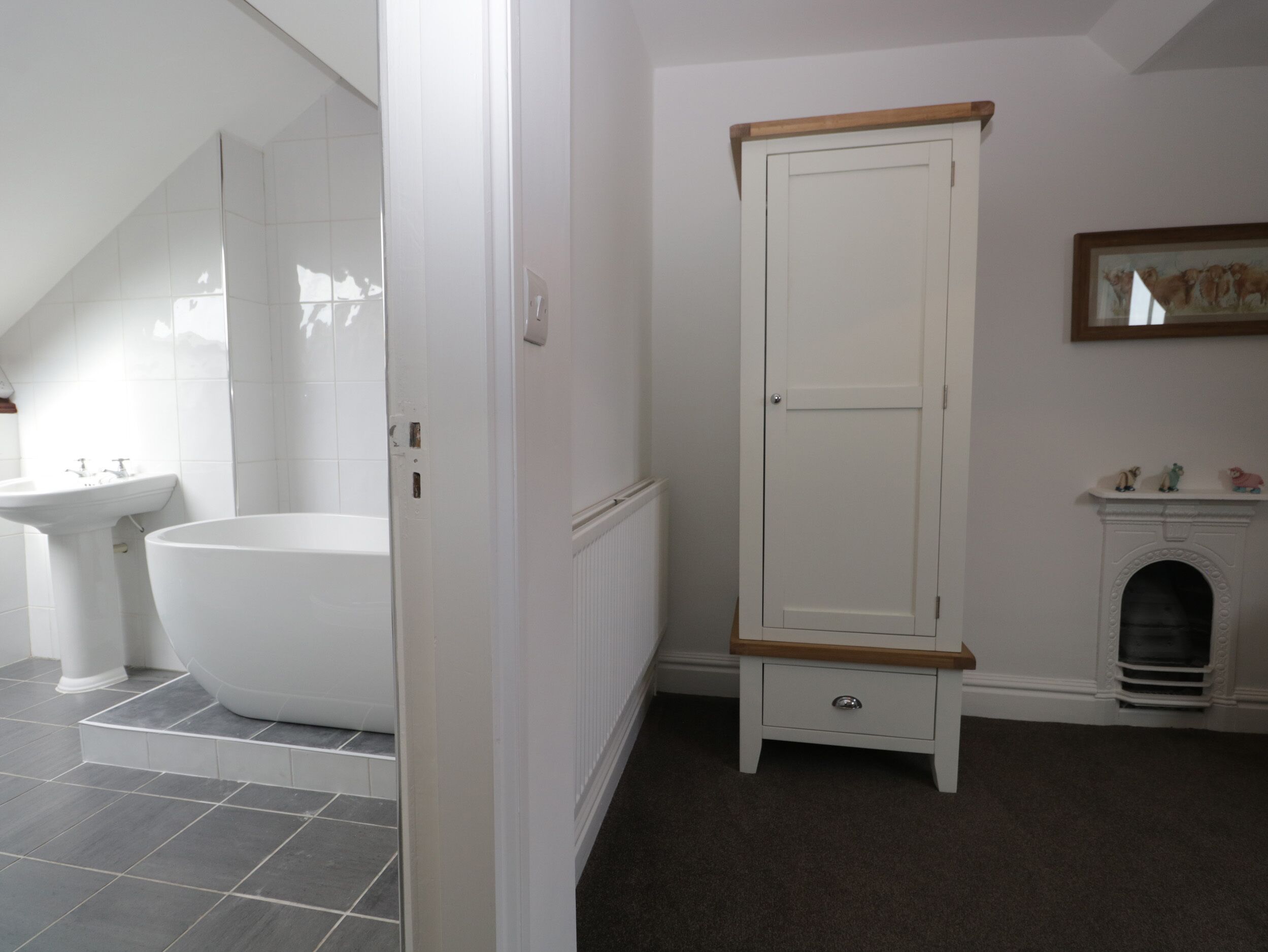 A bathroom with a bathtub, a white pedestal sink, a white wardrobe with a wooden top, and a fireplace with figurines.