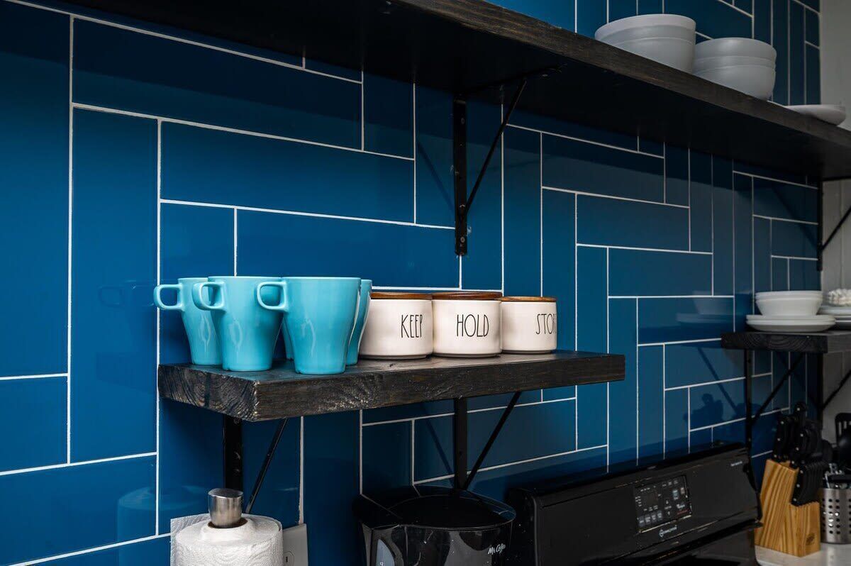 Eine Küche mit blauen Fliesen, einem Regal mit blauen Tassen und weißen Behältern sowie einer Kaffeemaschine.