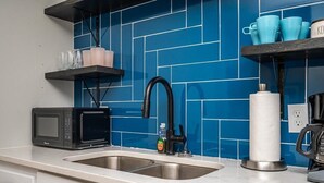 Una cocina con un salpicadero de azulejos azules, un microondas negro, un fregadero y una repisa con tazas azules y un tarro.