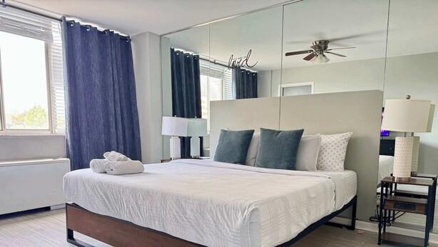 Una cama bien hecha con edredón blanco y almohadas, una mesita de noche con lámpara, un ventilador de techo y un espejo grande.