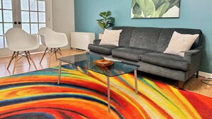 Una sala de estar con un sofá, dos sillas blancas, una mesa de centro de vidrio y un tapete colorido a rayas.