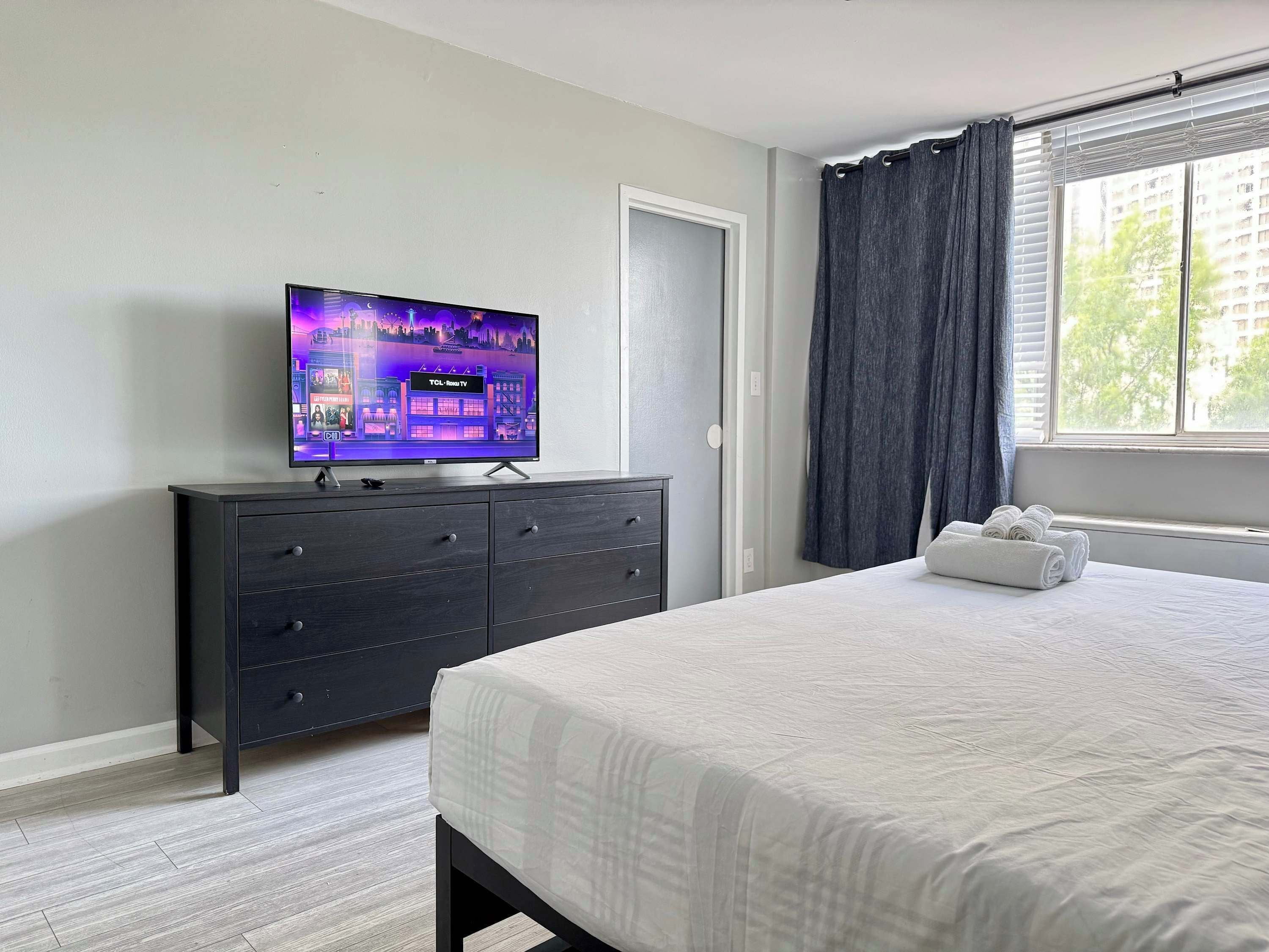 A hotel room with a bed, a TV on a dresser, a window with curtains, and a view of buildings outside.