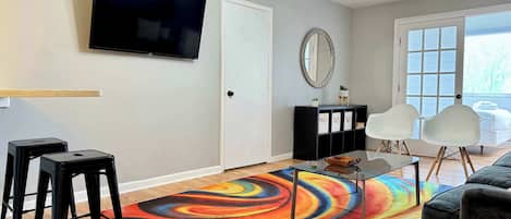 Una sala de estar con una televisión de pantalla plana, un tapete colorido, un mueble negro, sillas blancas y un sofá gris.