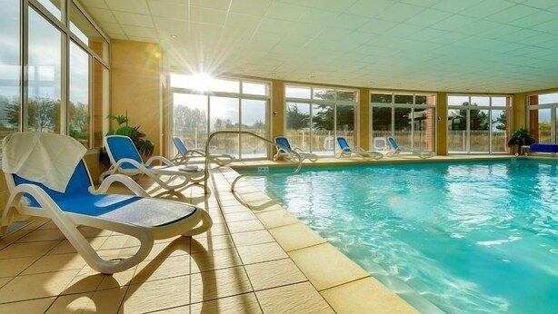 Una piscina interior con agua cristalina azul, rodeada de habitaciones iluminadas por el sol con ventanas amplias y sillones reclinables azules.