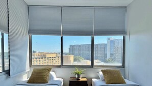 A hotel room with two beds, a small table, and a view of the cityscape through large windows.