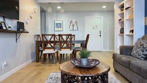 A living room with a dining area, a round coffee table with a plant, a grey sofa, and a TV mounted on the wall.