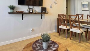 A dining area with a TV mounted on the wall, a wooden dining table with chairs, a woven placemat, and a potted plant.