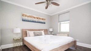 A bedroom with a wooden bed, two bedside lamps, a ceiling fan, a window, and a painting on the wall.