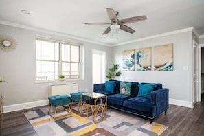 Un salon avec un canapé bleu, une table basse, un ventilateur de plafond et des œuvres d’art accrochées au mur.