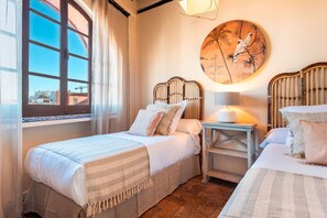 Une chambre avec deux lits, une fenêtre avec des rideaux, une lampe fixée au mur et un tableau rond.
