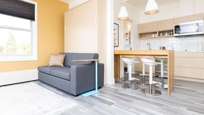 A modern living room with a grey sofa, a wooden dining table with bar stools, a kitchen area with a microwave, and a large window with blinds. - Clean, Central Plush Sanctuary in Highlands (Edmonton)