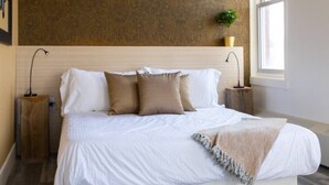 A neatly made bed with white and beige bedding, a wooden headboard, and a bedside table with a lamp. - Clean, Central Plush Sanctuary in Highlands (Edmonton)