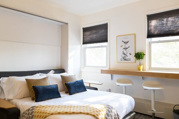 Een moderne slaapkamer met een groot bed, twee ramen met zonwering, een bureau met stoel, en een lijst met een afbeelding aan de muur.