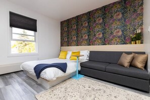 A bedroom with a bed, a grey sofa, a floral wall, a window with blinds, and a small table.