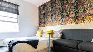 A bedroom with a bed, a grey sofa, a floral wallpaper, and a window with blinds. - Sparkling Clean, Central, Comfy & Quiet in Highlands (Edmonton)