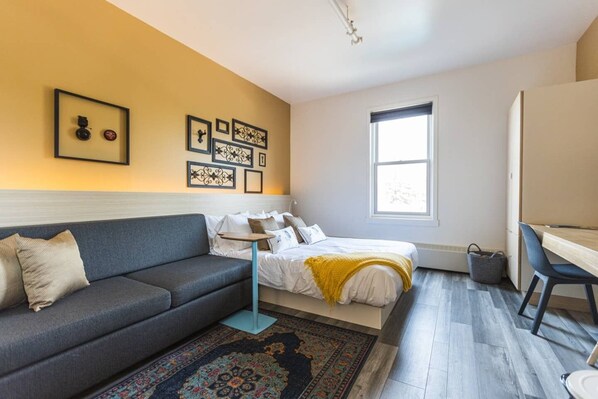 A modern bedroom with a grey sofa, a bed with a yellow throw, a desk, and a large window. - Clean Sophisticated Comfort in Highlands (Edmonton)