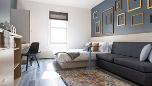 A modern bedroom with a bed, a grey sofa, a desk with a chair, and a window with blinds. - Clean, Charming & Cozy Sanctuary in Highlands (Edmonton)
