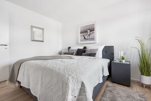 A modern bedroom with a large bed, a bedside table, a framed picture on the wall, and a potted plant. - Marina Sunrise - Harbor view, balcony, waterfront location, 2nd floor, parking (Olpenitz)