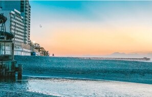 A coastal scene with a pier extending into the water, a building with a balcony overlooking the beach, and a clear sky.