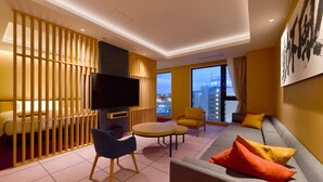 Quarto de hotel moderno com sofá, cadeiras, televisão e uma grande janela com vista para a cidade.