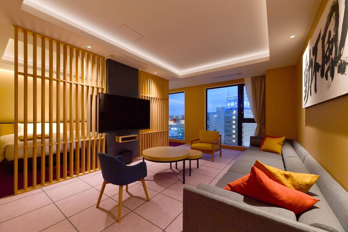 A modern hotel room with a sofa, chairs, a TV, and a large window offering a city view.