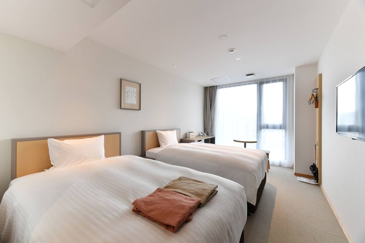 A hotel room with two beds, a desk, a chair, a TV, and large windows.