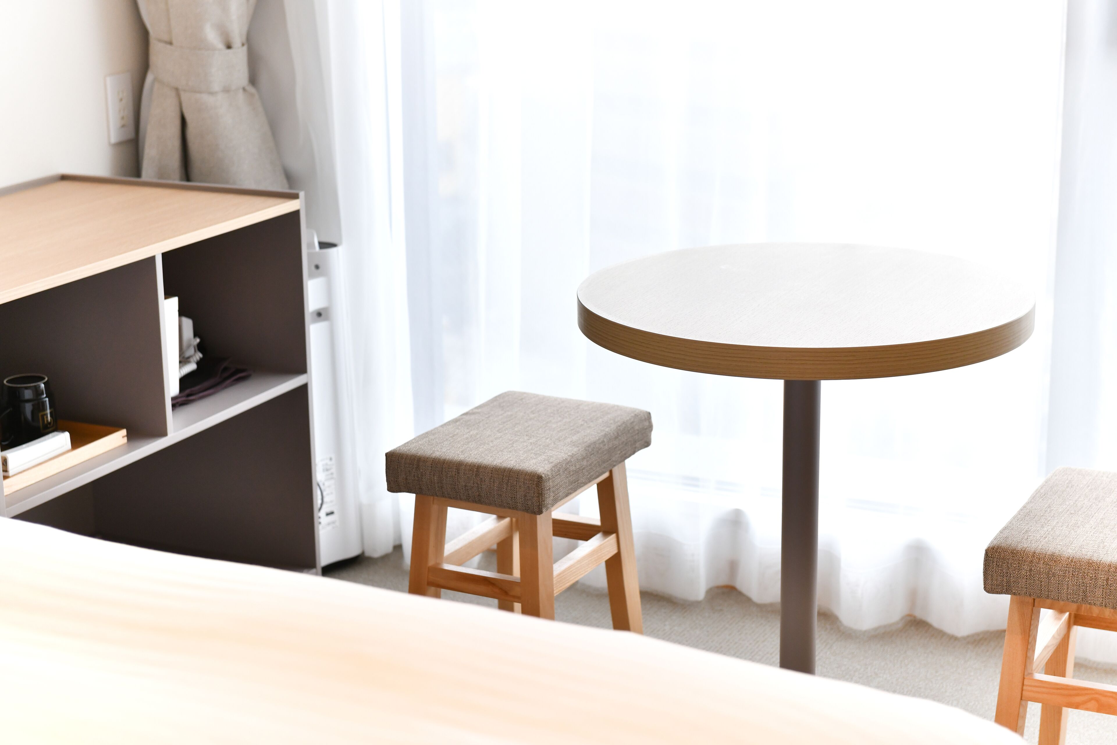 A modern room with a round table, two wooden stools, a bookshelf, and a bed.
