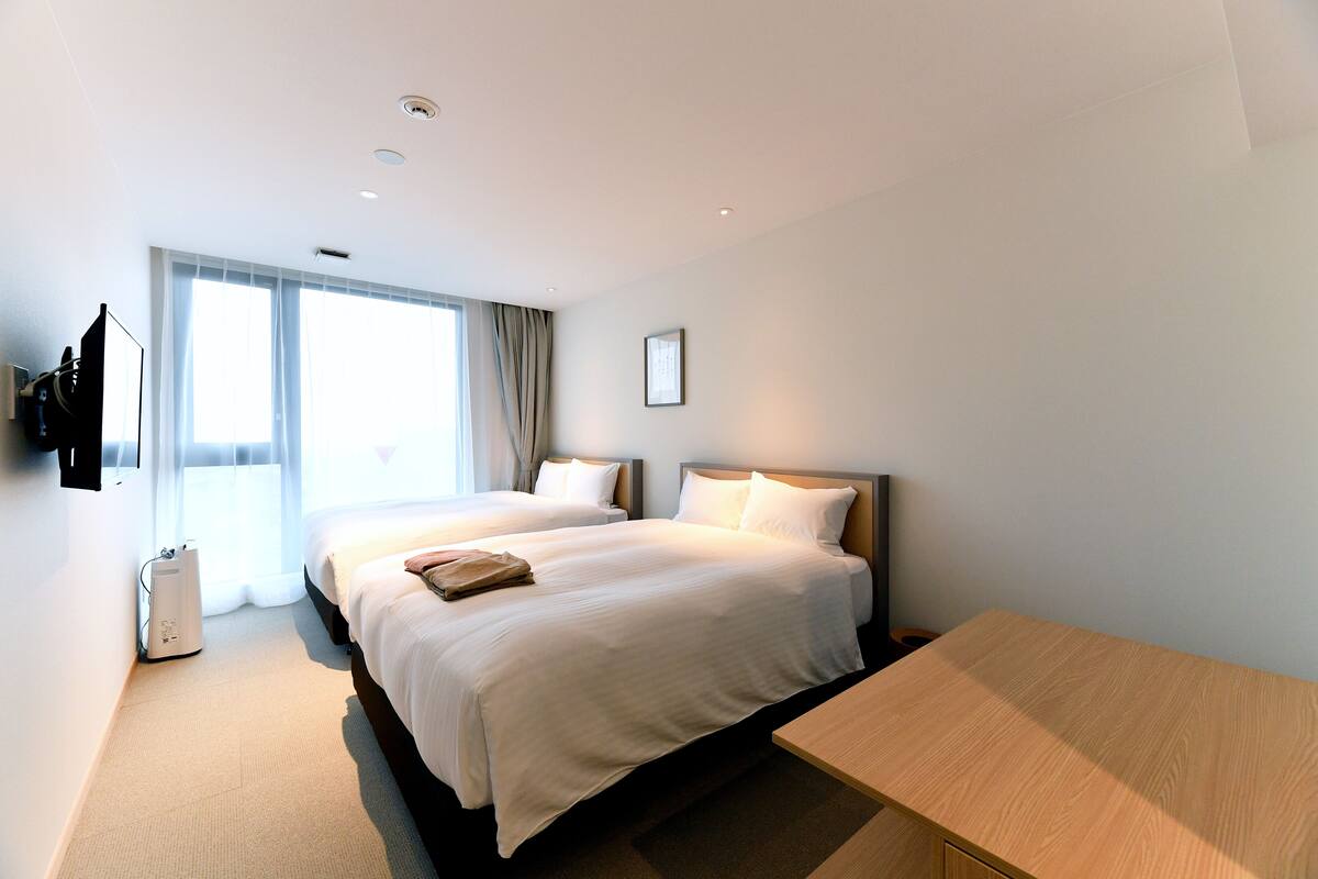 A modern hotel room with two beds, a wooden desk, and a large window with curtains.