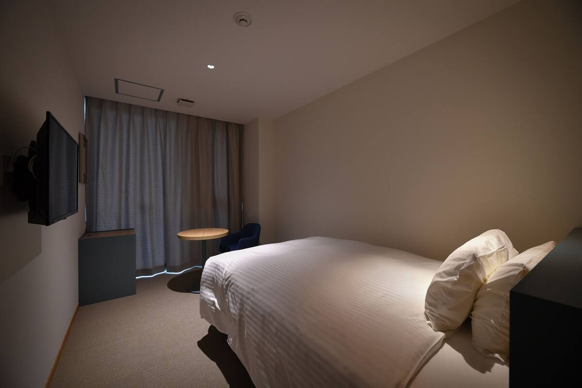 A hotel room with a bed, a desk, a chair, a flat-screen TV, and a window with curtains.
