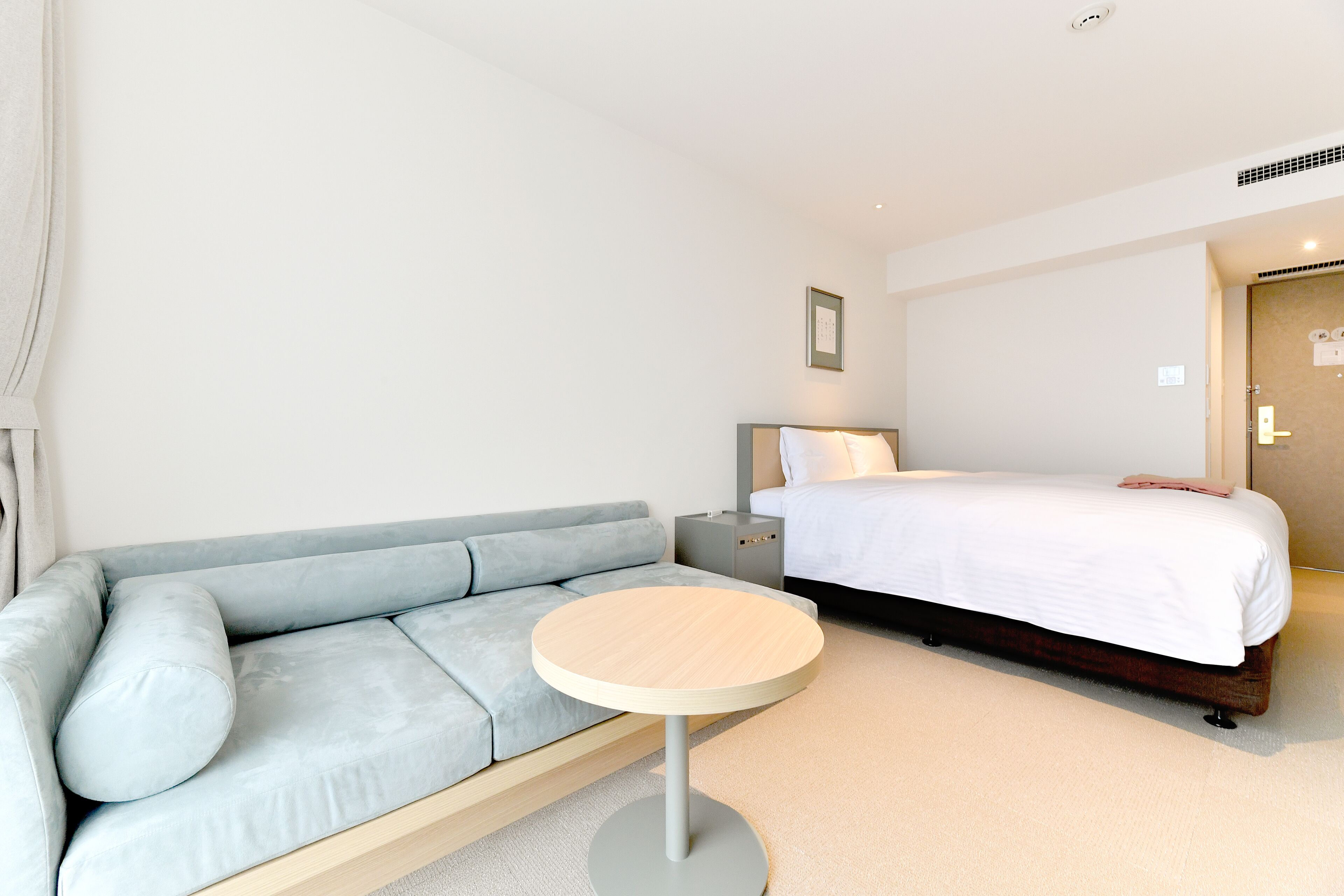 A modern hotel room with a light blue sofa, a round wooden table, a bed with white bedding, and a nightstand with a lamp.