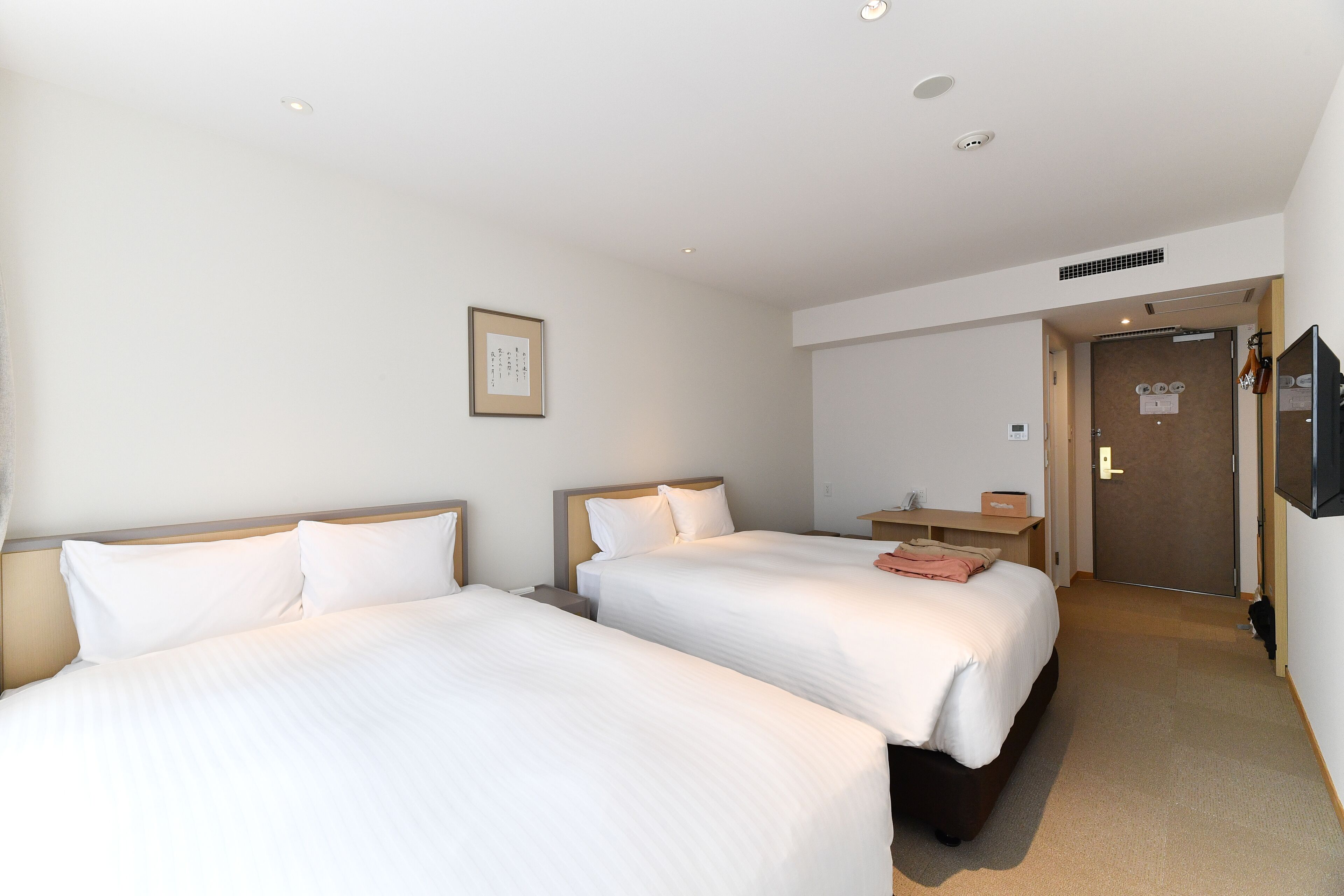 A hotel room with two beds, a wooden headboard, a bedside table, a TV mounted on the wall, and a door leading to a bathroom.