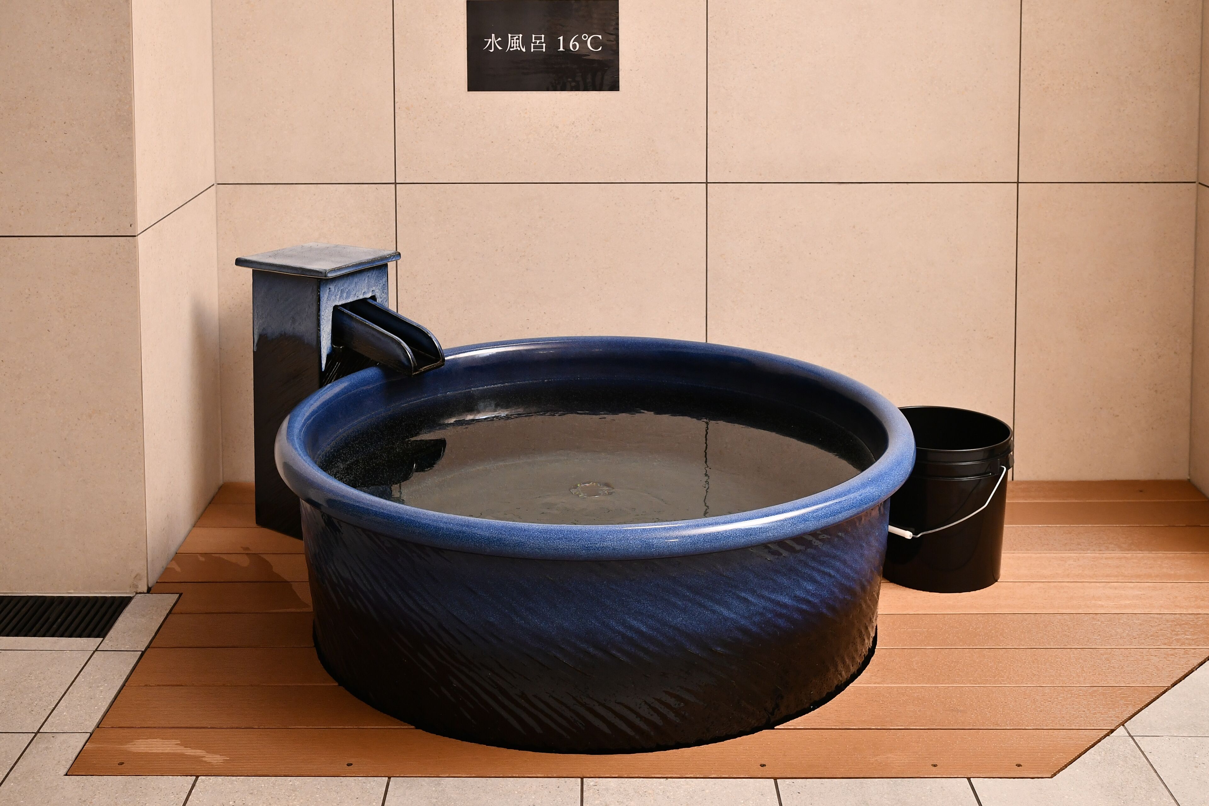 A blue hot tub with a black bucket beside it, placed on a wooden floor with a tiled wall in the background.