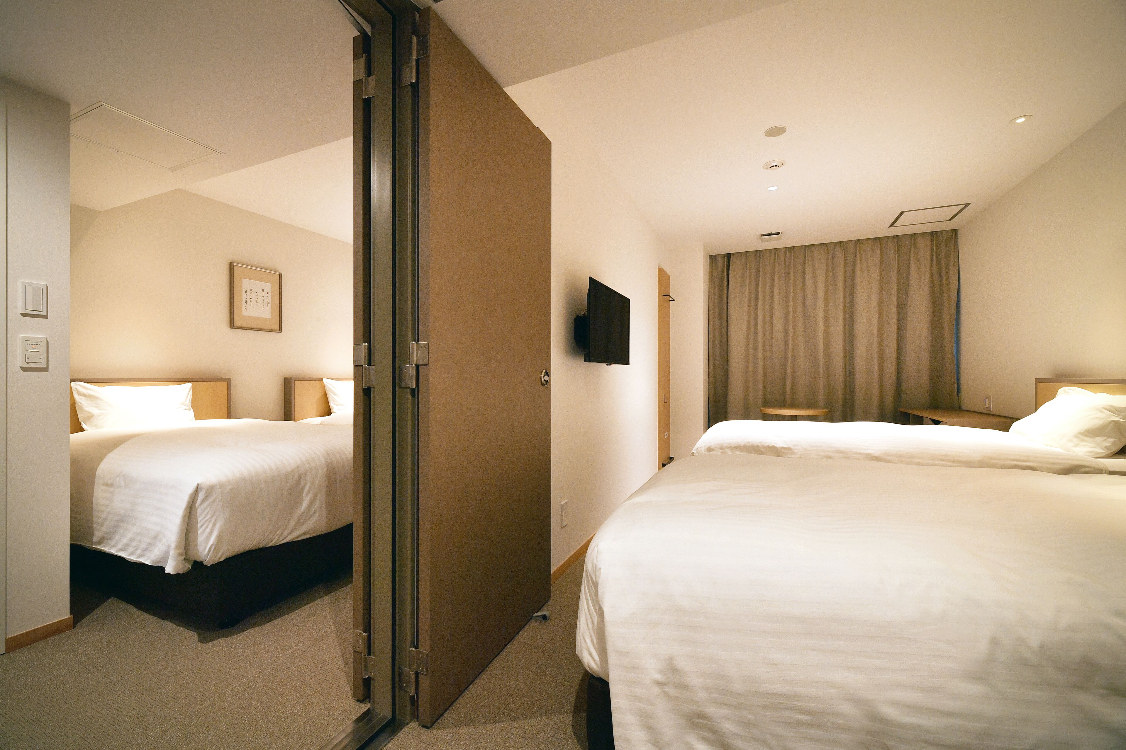 A hotel room with two beds, a flat-screen TV, a large window with curtains, and a door leading to another room.