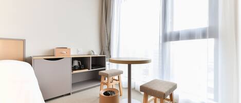 A hotel room with a bed, a desk, a chair, a small table, and a trash bin.