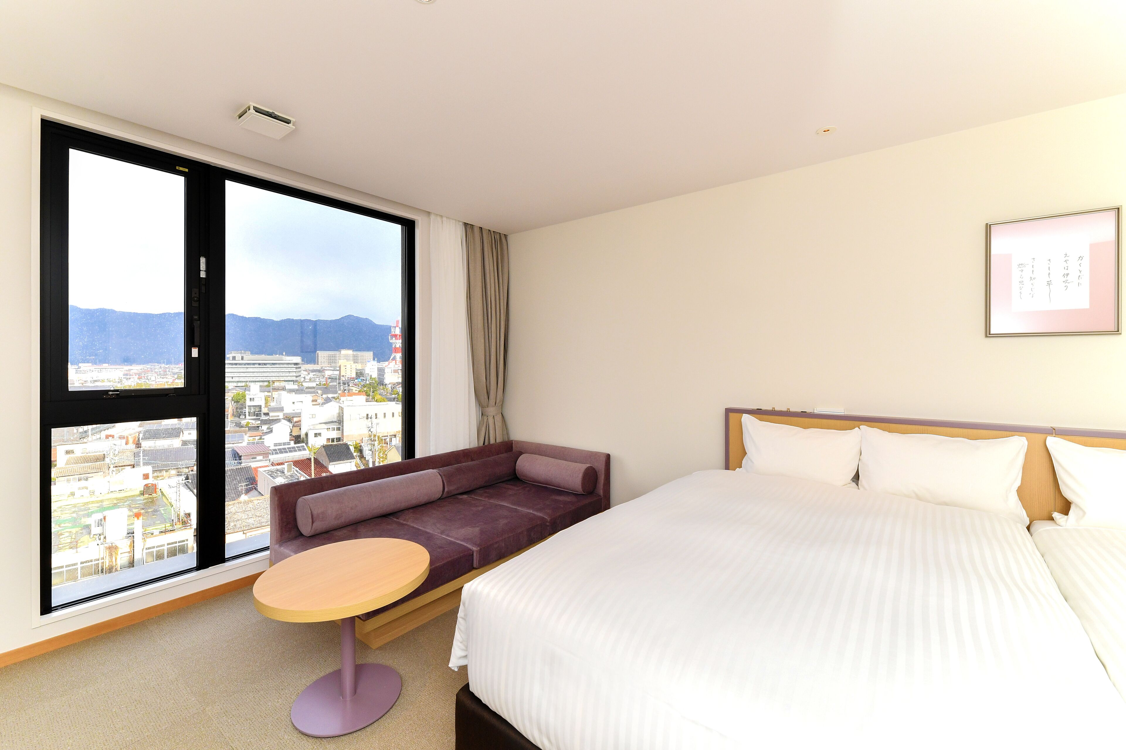 A hotel room with a large bed, a sofa bed, a small round table, and a large window offering a city view.