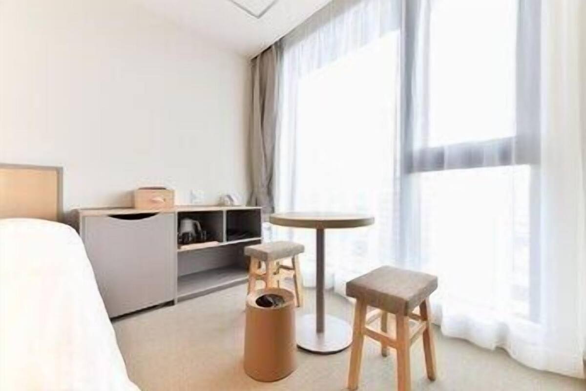 A hotel room with a bed, a small table, a stool, a trash bin, and a minibar.