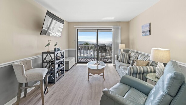 Ein Wohnzimmer mit einer Couch, einem Sessel und einem runden Couchtisch. An der Wand ist ein Fernseher angebracht und es gibt einen Balkon mit Meerblick.